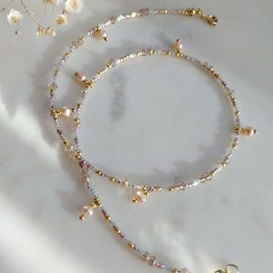 Könnte beinhalten: Zarte Perlenkette mit einem mehrschichtigen Design. Die Halskette enthält kleine goldene Perlen, hellrosa Perlen und klare und rosa Glasperlen. Die Halskette hat einen goldenen Verschluss. Der Schmuck wird auf einer weißen Oberfläche präsentiert.