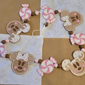 Op de afbeelding: Een slinger met peperkoek-stijl Mickey en Minnie Mouse hoofden en snoepornamenten. De ornamenten zijn lichtbruin, roze en wit, verbonden door houten kralen. Ideaal voor feestelijke decoraties, de slinger is een charmante toevoeging.