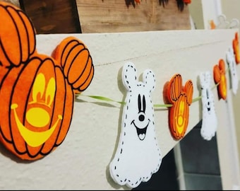 May include: A Halloween garland with orange and white felt decorations. The decorations are shaped like pumpkins and ghosts with Mickey Mouse ears. The garland is strung together with green string.