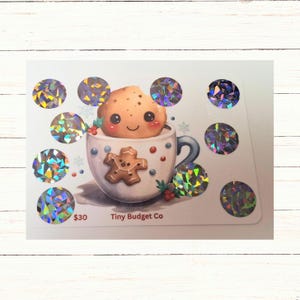 May include: A white card featuring a cartoon cookie in a mug with a gingerbread man design. The mug is decorated with snowflakes and berries. Surrounding the mug are several holographic stickers. The text "Tiny Budget Co" is visible.