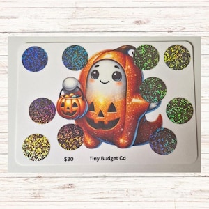 May include: A white card featuring a cartoon ghost in a pumpkin costume holding a jack-o'-lantern bucket. The card has ten glittery circles in various colors. Text on the card reads "$30 Tiny Budget Co."