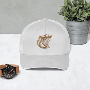 May include: White trucker hat with a brown embroidered image of a squirrel wearing a cowboy hat.