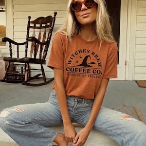 May include: A rust-colored short-sleeve t-shirt featuring a black graphic print that reads Witches Brew Coffee Co. Stirring Up Trouble, accompanied by a witch hat, moon, and bat icons, paired with light blue distressed denim jeans.