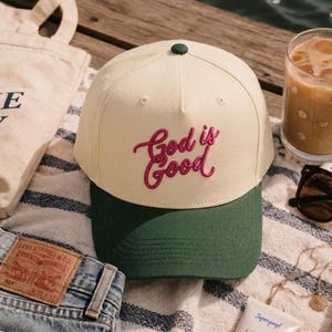 Op de afbeelding: Een baseballpet met een crèmekleurige kroon en een donkergroene klep. De voorkant van de pet is voorzien van de geborduurde tekst "God is Good" in een donkerroze cursief lettertype. De pet heeft een kleine donkergroene knoop bovenop.