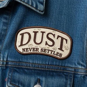 May include: Close-up of a blue denim jacket with a patch that reads "DUST NEVER SETTLES" in brown and cream. The jacket has silver buttons and two chest pockets.