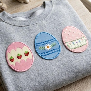 Può includere: Una felpa grigia con tre toppe ricamate a forma di uovo di Pasqua. Le toppe sono rosa, blu e rosa, ognuna con disegni unici tra cui fragole, motivi floreali e pois. Un coniglietto di ceramica bianca e rametti decorativi sullo sfondo.