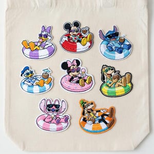 May include: A beige tote bag featuring embroidered patches of Disney characters relaxing in inner tubes. Characters include Mickey Mouse, Minnie Mouse, Donald Duck, Daisy Duck, Stitch, and Goofy, all wearing sunglasses and holding drinks.