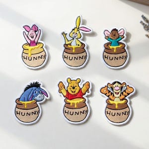 May include: Embroidered patches featuring characters from Winnie the Pooh, including Piglet, Rabbit, and Eeyore, each emerging from a honey pot. The patches have a white border and the word "HUNNY" is printed on the pot.