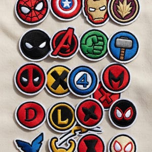 May include: A collection of round, embroidered patches featuring various superhero logos and symbols. The patches showcase vibrant colors and detailed designs, including Spider-Man, Captain America, and Iron Man. These patches are likely for clothing or accessories.
