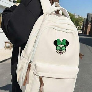 May include: Off-white backpack with a front pocket and brown zipper pulls. The backpack features a Minnie Mouse patch with a green bow and sunglasses. The bag has a top handle and adjustable shoulder straps.