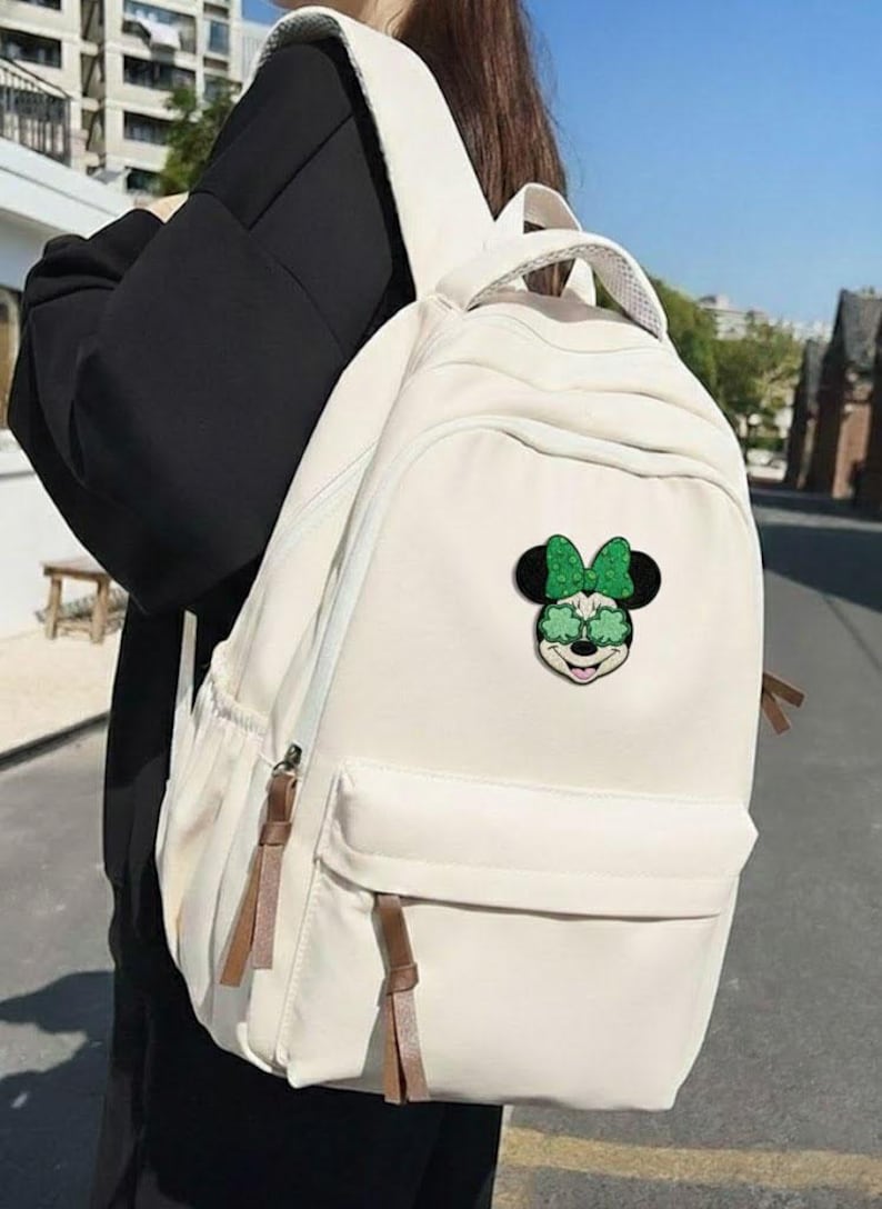 May include: Off-white backpack with a front pocket and brown zipper pulls. The backpack features a Minnie Mouse patch with a green bow and sunglasses. The bag has a top handle and adjustable shoulder straps.
