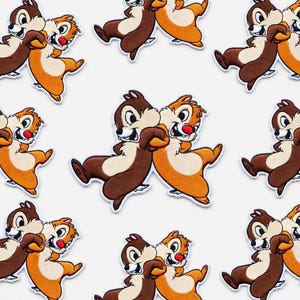 Könnte beinhalten: Ein Muster aus gestickten Aufnähern mit Chip und Chap, den Disney-Chipmunks. Jeder Aufnäher zeigt die Charaktere in einer spielerischen Pose, mit braunem und orangefarbenem Fell, weißen Akzenten und roten Nasen. Auf weißem Hintergrund.