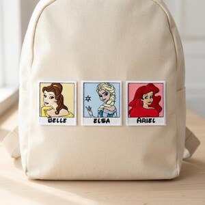 May include: Beige backpack featuring embroidered patches of Belle, Elsa, and Ariel. Each patch is in a polaroid-style frame with the character's name below. The backpack has adjustable straps and a top handle.