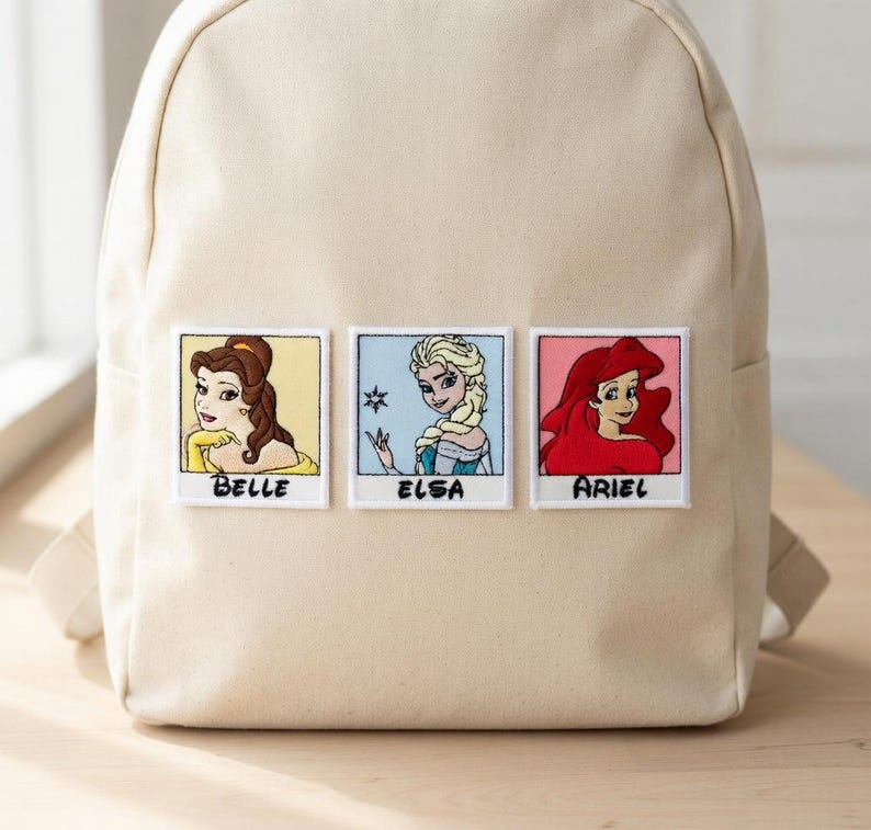 May include: Beige backpack featuring embroidered patches of Belle, Elsa, and Ariel. Each patch is in a polaroid-style frame with the character's name below. The backpack has adjustable straps and a top handle.