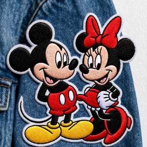 Könnte beinhalten: Gestickte Aufnäher von Mickey Mouse und Minnie Mouse auf einer Jeansjacke. Mickey trägt rote Shorts, gelbe Schuhe und einen schwarzen Körper. Minnie trägt ein rotes Kleid und eine Schleife mit weißen Handschuhen. Die Aufnäher sind weiß umrandet.