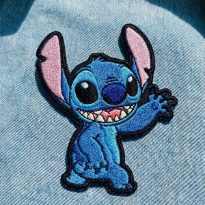 May include: Embroidered patch of the character Stitch from Lilo & Stitch. The patch is blue with black outlines, pink inner ears, and a smiling expression. The patch is designed to be ironed or sewn onto fabric.