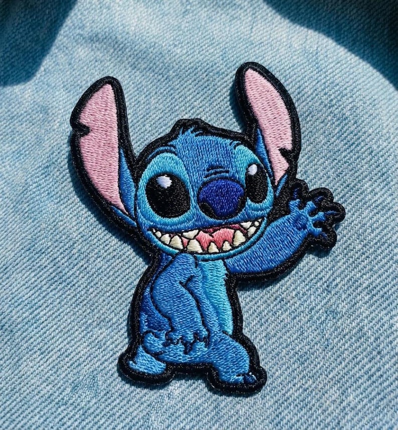 May include: Embroidered patch of the character Stitch from Lilo & Stitch. The patch is blue with black outlines, pink inner ears, and a smiling expression. The patch is designed to be ironed or sewn onto fabric.