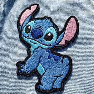 May include: Embroidered patch of the blue alien Stitch from Lilo & Stitch. The patch has a black border, pink inner ears, and is shown on a light blue denim background. The character is in a playful pose.