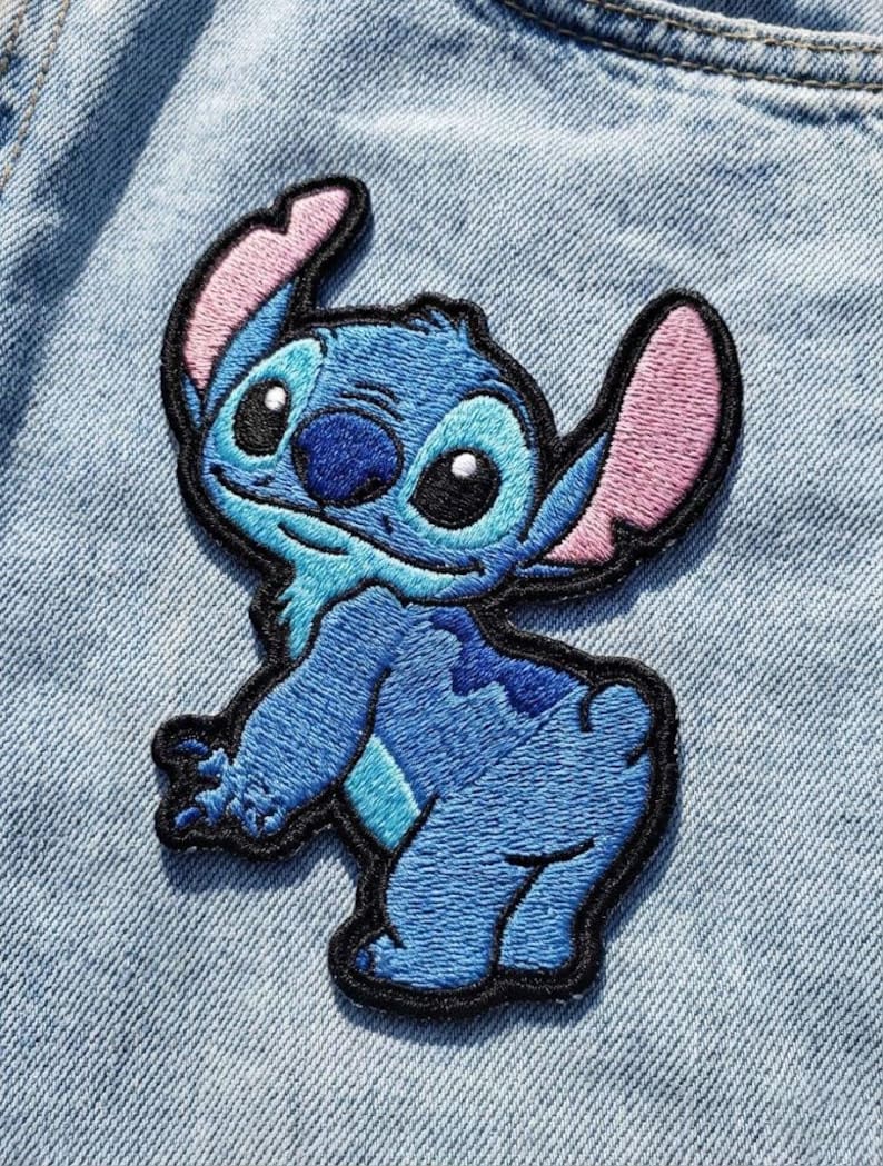 May include: Embroidered patch of the blue alien Stitch from Lilo & Stitch. The patch has a black border, pink inner ears, and is shown on a light blue denim background. The character is in a playful pose.