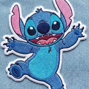 May include: Embroidered patch of the character Stitch from Lilo & Stitch. The patch shows Stitch in various shades of blue, with pink ears and a white border. The design depicts Stitch with a wide grin and outstretched arms.