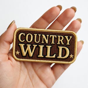 May include: A rectangular patch with the words "COUNTRY WILD" in gold lettering on a brown background. The patch has a gold border and two small gold stars. The patch is held in a hand.