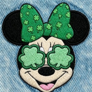 May include: Embroidered patch featuring a cartoon mouse with a green bow adorned with shamrocks. The mouse's eyes are replaced with green shamrock shapes. The patch is set against a light blue denim background.