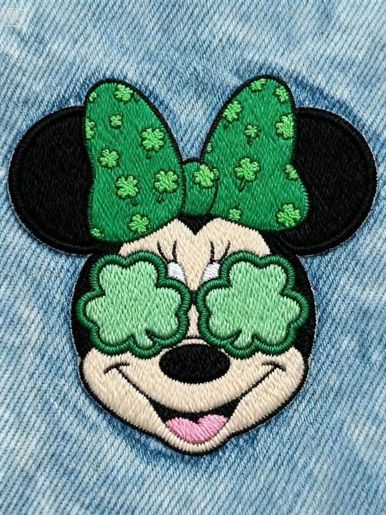 May include: Embroidered patch featuring a cartoon mouse with a green bow adorned with shamrocks. The mouse's eyes are replaced with green shamrock shapes. The patch is set against a light blue denim background.