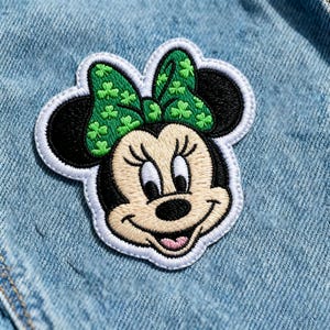 Può includere: Toppa ricamata con Minnie Mouse con un fiocco verde decorato con trifogli. La toppa ha un bordo bianco ed è posta su uno sfondo di denim blu chiaro. Il design è dettagliato e colorato.