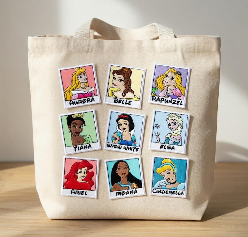 May include: Beige canvas tote bag with nine embroidered patches featuring Disney princesses: Aurora, Belle, Rapunzel, Tiana, Snow White, Elsa, Ariel, Moana, and Cinderella. The patches are rectangular with white borders.