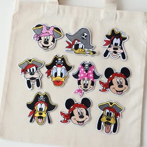 Peut inclure: Plusieurs écussons de personnages de dessins animés, dont Mickey Mouse, Minnie Mouse, Donald Duck et Dingo, tous déguisés en pirates. Les écussons présentent des chapeaux de pirates noirs, des bandanas rouges et des détails de têtes de mort. Sur un sac fourre-tout beige.