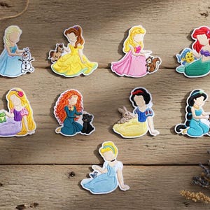Könnte beinhalten: Eine Sammlung farbenfroher, gestickter Aufnäher mit verschiedenen Prinzessinnen-Charakteren. Jeder Aufnäher zeigt eine Prinzessin in einem detaillierten Kleid, oft mit einem tierischen Begleiter, vor einem Holzgrund. Die Aufnäher sind in verschiedenen Farben.