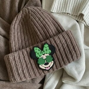 May include: A brown knit beanie with a folded brim. The beanie features a patch of Minnie Mouse wearing green shamrock sunglasses and a matching bow. The hat is made of a ribbed knit material.