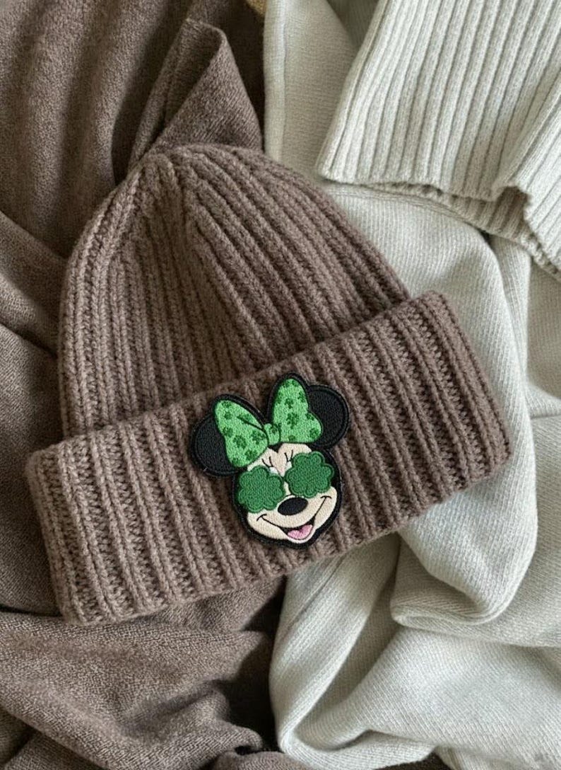 May include: A brown knit beanie with a folded brim. The beanie features a patch of Minnie Mouse wearing green shamrock sunglasses and a matching bow. The hat is made of a ribbed knit material.