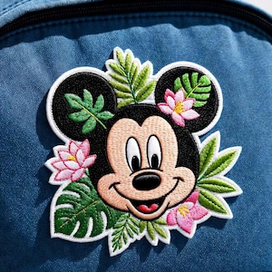 Puede incluir: Parche bordado con Mickey Mouse con un diseño floral tropical. El parche tiene un contorno negro, con la cara de Mickey en color beige y negro. Flores de loto rosas y hojas verdes rodean al personaje, sobre un fondo de mezclilla azul.