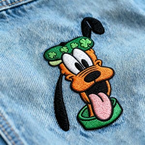 May include: Embroidered patch of a cartoon dog with orange fur, white eyes, and a pink tongue. The dog wears a green hat with shamrocks. The patch is sewn onto light blue denim fabric.