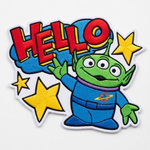May include: Embroidered patch featuring a green three-eyed alien character in a blue jumpsuit, waving with the word "HELLO" in red letters above. The design includes yellow stars and a blue cloud background.