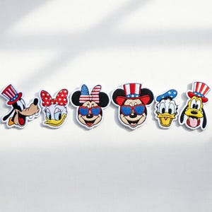 以下が含まれることがあります： 愛国的な衣装を着た漫画のキャラクターが特徴の刺繍パッチ。 グーフィー、デイジーダック、ミニーマウス、ミッキーマウス、ドナルドダック、プルートが含まれており、それぞれに赤、白、青のアクセントがあり、衣類やアクセサリーに最適です。