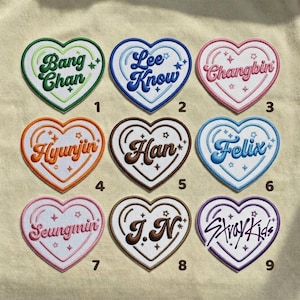 Peut inclure: Neuf écussons brodés en forme de cœur avec des noms et le texte "Stray Kids". Chaque écusson a une palette de couleurs différente, dont le vert, le bleu, le rose, l'orange, le marron et le violet. Les écussons sont sur un fond beige.