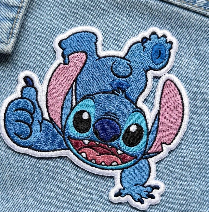 May include: Embroidered patch of the character Stitch from Lilo & Stitch. The patch depicts Stitch in a playful pose, with blue and pink colours, and a white border. The patch is on a denim background.
