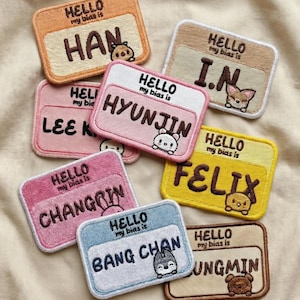 May include: Embroidered name tag patches in various colors, featuring names like "HAN," "HYUNJIN," and "FELIX." Each patch includes the phrase "HELLO my bias is" and a small cartoon animal. The patches are rectangular with rounded corners.