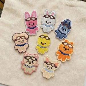 May include: A collection of eight embroidered patches featuring various animal characters, including a pig, rabbit, dog, duck, fox, and cat. Each patch is adorned with aviator goggles and a bow tie or collar, in a variety of colors.