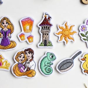 Op de afbeelding: Geborduurde patches met personages en elementen uit een sprookje. Inclusief een prinses in een paarse jurk, een toren, een zon, bloemen, een kroon, een stel, een kameleon, een koekenpan, een eend en een zwevende lantaarn.