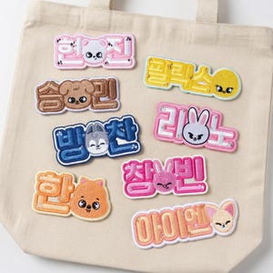 Puede incluir: Colección de parches bordados con texto coreano y caras de animales lindos. Los parches son de varios colores, incluyendo rosa, amarillo, naranja y azul, y están adheridos a una bolsa de mano beige. Cada parche presenta un diseño de animal diferente.