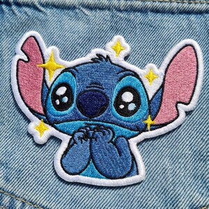 May include: Embroidered patch of the character Stitch from Lilo & Stitch. The patch has a white border, blue body, pink ears, and large black eyes. Yellow star accents are around the character's head.