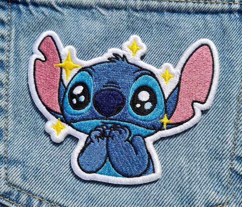 May include: Embroidered patch of the character Stitch from Lilo & Stitch. The patch has a white border, blue body, pink ears, and large black eyes. Yellow star accents are around the character's head.