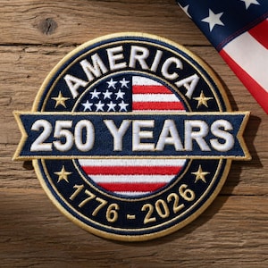 Op de afbeelding: Geborduurde patch met de tekst "AMERICA 250 YEARS 1776-2026" in wit en goud op een marineblauwe achtergrond. De patch toont de Amerikaanse vlag en sterren. Een Amerikaanse vlag staat op de achtergrond.