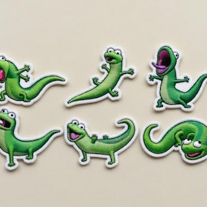 Peut inclure: Ensemble de six écussons brodés représentant des lézards verts avec des bordures blanches. Chaque écusson présente une pose différente, avec des bouches ouvertes et des détails expressifs. Parfait pour ajouter une touche amusante et fantaisiste aux vêtements ou accessoires.