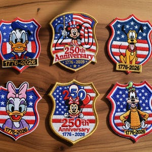 May include: A selection of embroidered patches showcasing Disney characters in patriotic shield designs. Each patch features a red, white, and blue colour scheme with stars and stripes, and the text "1776-2026". Characters include Donald Duck, Mickey Mouse, Pluto, Daisy Duck, Minnie Mouse, and Goofy.