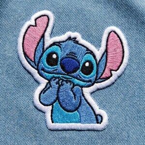 May include: Embroidered patch of the character Stitch from Lilo & Stitch. The patch is blue with pink ears, black eyes, and a white border. The character is in a pose with its hands clasped together.