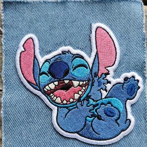 May include: Embroidered patch of the character Stitch from Lilo & Stitch. The patch shows Stitch in blue, with pink inner ears and mouth, and a white outline. The patch is set on a denim background.
